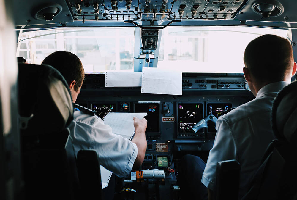 Two airline pilots seated in a flight deck, with one reviewing flight paperwork.