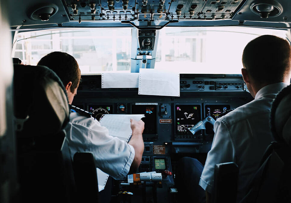 Two airline pilots seated in a flight deck, with one reviewing flight paperwork.